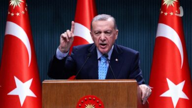 رجب طیب اردوغان رئیس‌جمهور ترکیه در حال سخنرانی با انگشت اشاره رو به بالا پشت تریبون رسمی و میان دو پرچم ترکیه.