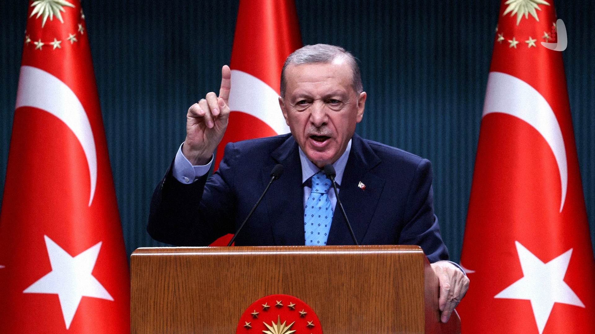 رجب طیب اردوغان رئیس‌جمهور ترکیه در حال سخنرانی با انگشت اشاره رو به بالا پشت تریبون رسمی و میان دو پرچم ترکیه.