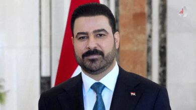 محمد شیاع السودانی نخستوزیر عراق با کت و شلوار تیره، کراوات آبیرنگ و پیکسل پرچم عراق روی یقه کت.