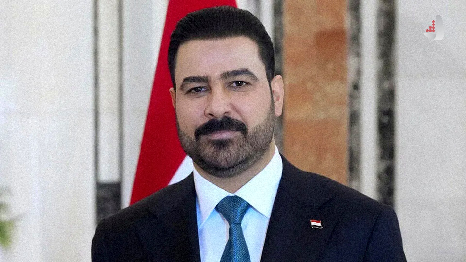 محمد شیاع السودانی نخست‌وزیر عراق با کت و شلوار تیره، کراوات آبی‌رنگ و پیکسل پرچم عراق روی یقه کت.