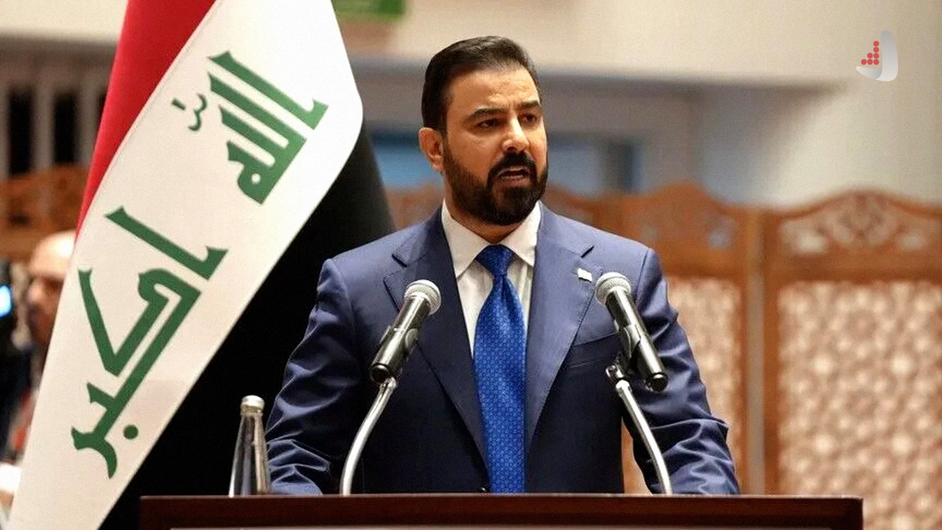 نمای روبرو از محمود المشهدانی رئیس پارلمان عراق با کت و شلوار سورمه‌ای و کراوات آبی در حال سخنرانی پشت تریبون در کنار پرچم عراق