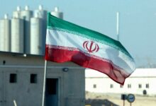 پرچم جمهوری اسلامی ایران در حال اهتزاز در محوطه یک تاسیسات صنعتی یا هسته‌ای با ساختمان‌ها و سازه‌های استوانه‌ای در پس‌زمینه