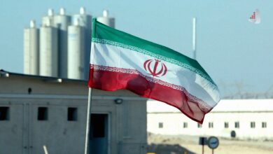 پرچم جمهوری اسلامی ایران در حال اهتزاز در محوطه یک تاسیسات صنعتی یا هسته‌ای با ساختمان‌ها و سازه‌های استوانه‌ای در پس‌زمینه