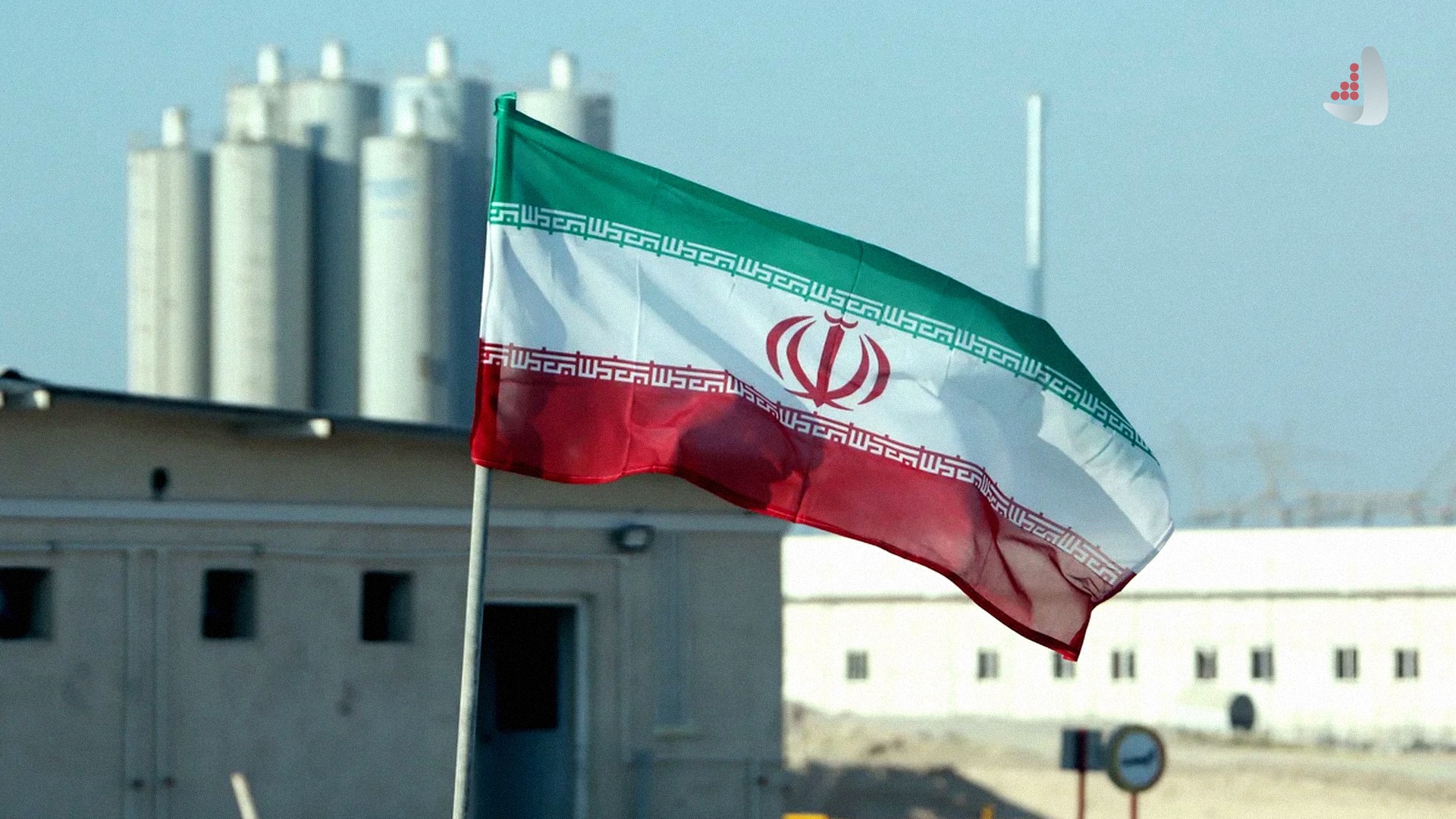 پرچم جمهوری اسلامی ایران در حال اهتزاز در محوطه یک تاسیسات صنعتی یا هسته‌ای با ساختمان‌ها و سازه‌های استوانه‌ای در پس‌زمینه