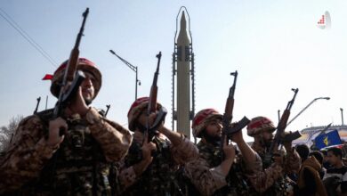 نیروهای نظامی با لباس استتار بیابانی و اسلحه کلاشنیکف در صفوف منظم همراه با یک موشک بالستیک در پس‌زمینه